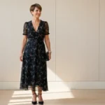 Elegancka kobieta w czarnej sukience midi na weselu, z biżuterią i szpilkami, patrząca do boku z uśmiechem